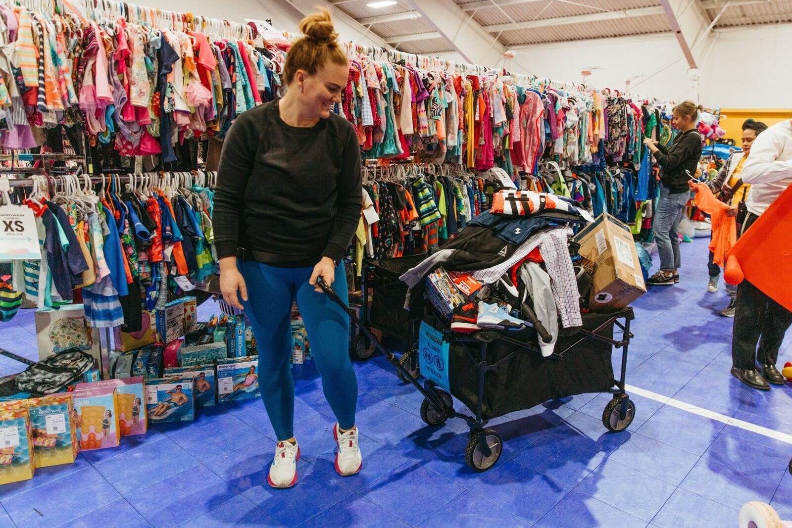 One smiling woman shopping in a clothing aisle pulling a wagon full of clothing, toys.